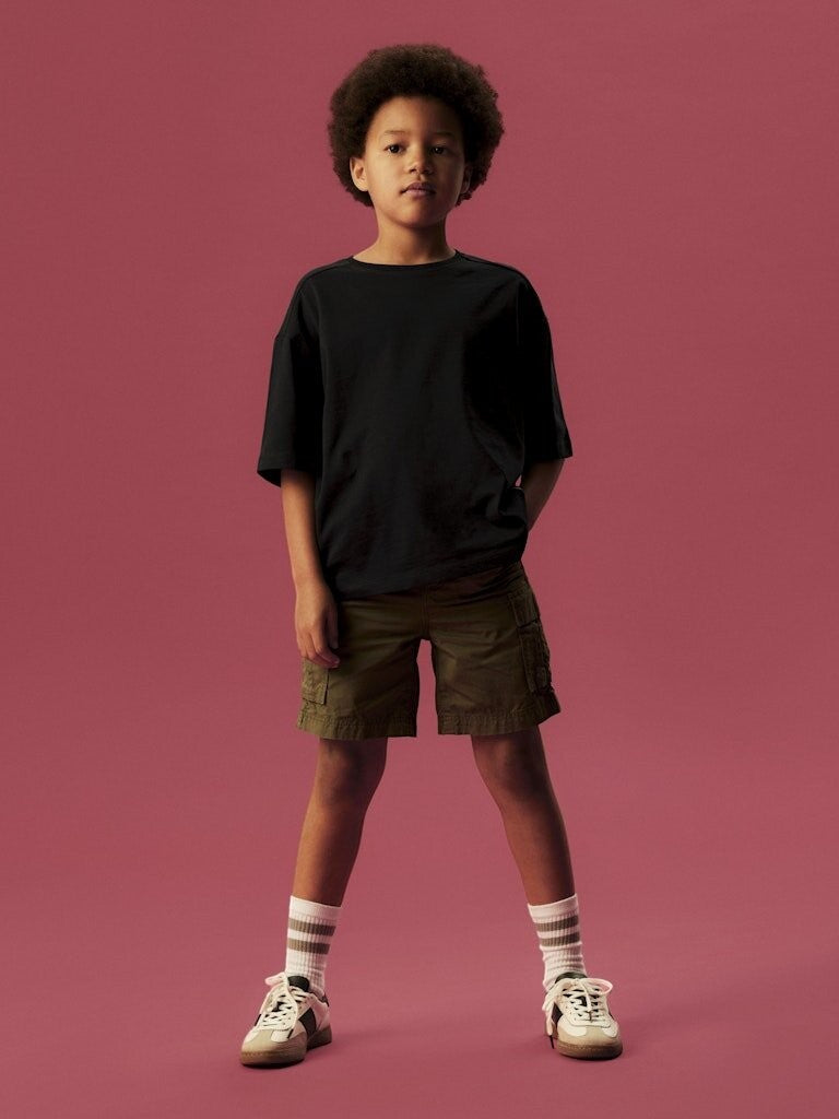 Bambino indossa bermuda cargo color olivine in cotone di Scugnizzi 0-16 con t-shirt nera e sneakers